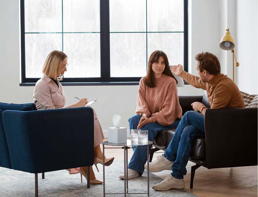 Woman and man having a serious conversation on a couch while a therapist takes notes in a bright room.