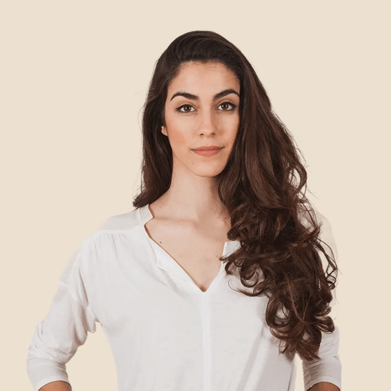 Confident woman with long curly brown hair wearing a white blouse standing against a beige background.